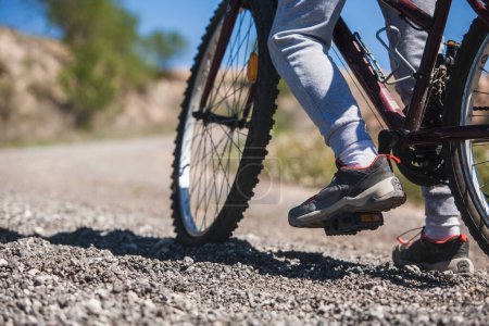 A person on a bicycle is standing with his foot on the ground on a rocky road. The person is wearing sneakers and socks.