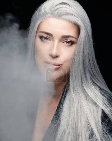 Foto de Retrato de moda de mujer modelo de pelo plateado soplando una gran nube de humo. Hermoso fumador aislado sobre fondo negro. Formato vertical - Imagen libre de derechos