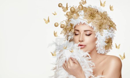 Photo for Beautiful model girl with golden festive makeup and hairstyle surrounded by golden butterflies, and dainty white feathers. High fashion portrait of a fairy concept for new year celebration over white - Royalty Free Image