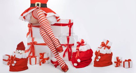 Foto de Mujer sexy de Navidad. Hermosa señora santa claus piernas en medias a rayas y tacones altos junto a regalos de Navidad y feliz inscripción de Navidad - Imagen libre de derechos