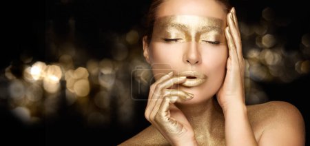 Téléchargez les photos : Fille modèle de beauté avec un maquillage doré métallique brillant tenant une main sur la joue tout en touchant les lèvres avec l'autre avec les yeux fermés dans un geste sensuel. Portrait de beauté sur fond bokeh - en image libre de droit