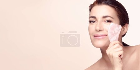 Téléchargez les photos : Beauty and skincare concept with a beautiful woman doing skin care routine with gua sha tool. Middle aged female using guasha. Massage for facial lifting. Beige background panorama banner. - en image libre de droit