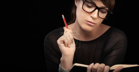 Foto de Close up Beautiful College Girl with Eyeglasses Writing on her Notebook. Isolated on Black Background with Copy Space - Imagen libre de derechos