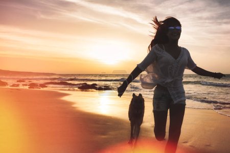 Téléchargez les photos : Libérer le bonheur. Femme et chien qui courent sans soucis sur les rivages sablonneux au coucher du soleil, avec éblouissement intentionnel du soleil et éclat de lentille. - en image libre de droit