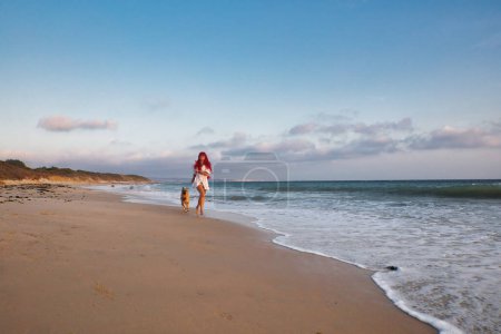 Photo for Red-haired woman and her dog running free along the beach at sunset. Relax, fun, and enjoy summer vacation - Royalty Free Image