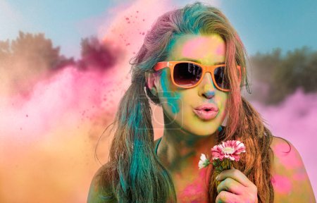 Téléchargez les photos : Belle femme sensuelle couverte de poudre colorée arc-en-ciel utilisée pour célébrer la fête des couleurs portant des lunettes de soleil colorées et soufflant doucement une fleur dans un concept de printemps de beauté - en image libre de droit