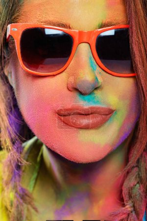 Foto de Hermosa mujer joven con gafas de sol cubiertas de polvo Holi de color arco iris. Retrato de estudio. Formato vertical - Imagen libre de derechos