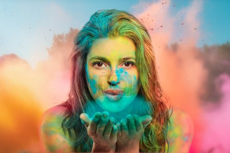 Photo for Young woman blowing holi powder from hands to camera. Happy woman covered in rainbow colored powder celebrating the festival of colors - Royalty Free Image