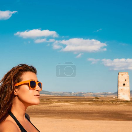 Photo for Beautiful woman in trendy colorful modern sunglasses walking along a deserted beach with historic stone tower in the background on a sunny summer day with copy space - Royalty Free Image