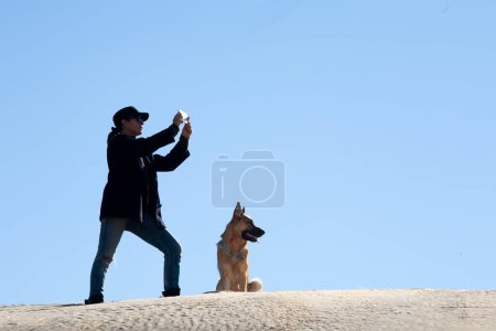 Photo for Woman with dog on sand dune under blue sky, taking photographs with a smartphone. Copy space - Royalty Free Image