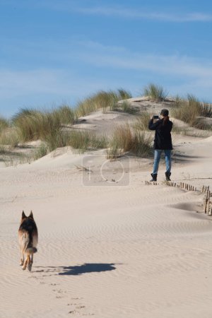 Foto de Una mujer captura la naturaleza con su smartphone en una duna de arena, acompañada por un perro. Con espacio de copia - Imagen libre de derechos