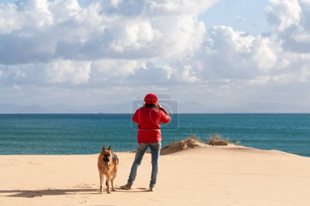 Téléchargez les photos : Femme et son chien marchant sur une plage de sable fin avec un ciel magnifique et une mer calme. Avec espace de copie - en image libre de droit