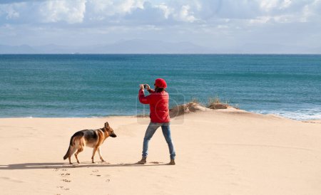 Photo for A woman stands on the beach, capturing the joy of a beloved pet against the backdrop of a wild and vibrant natural environment with her smartphone. - Royalty Free Image
