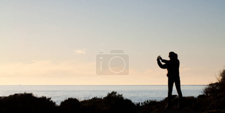 Téléchargez les photos : Capturer la beauté tranquille du lever du soleil sur la plage. Une femme debout, photographiant le paysage côtier serein de Tarifa en Espagne. Avec espace de copie - en image libre de droit