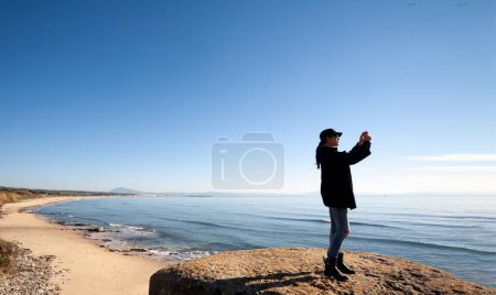 Foto de Una mujer captura la belleza costera con su cámara en una playa de arena, con cielos despejados y el vasto océano como telón de fondo en Tarifa Cádiz. Con copyspace - Imagen libre de derechos