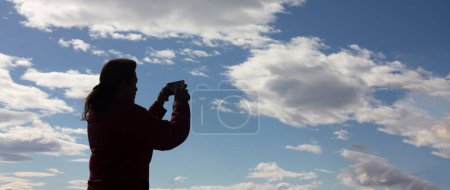 Téléchargez les photos : Une femme photographie le ciel avec un smartphone, capturant le fond nuageux. Avec espace de copie. - en image libre de droit