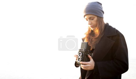 Foto de Joven fotógrafa sosteniendo cámara vintage, usando ropa casual y sombrero sobre fondo blanco. Con copyspace para añadir texto o gráficos. - Imagen libre de derechos