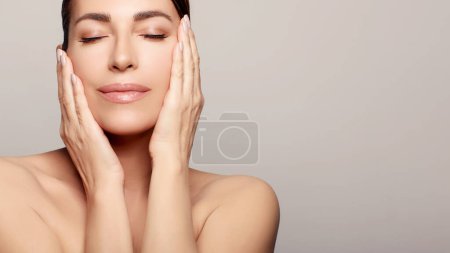 Photo for A beautiful woman with brown hair, eyes closed, relaxing indoors. Her hand gently lifts her cheek, emphasizing youthful skin. A serene spa-like image for skincare and rejuvenation. - Royalty Free Image