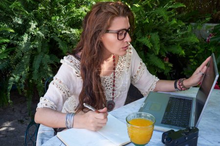 Photo for Young woman with long brown hair works and studies outdoors on her laptop. She wears glasses and enjoys a drink while communicating digitally. - Royalty Free Image