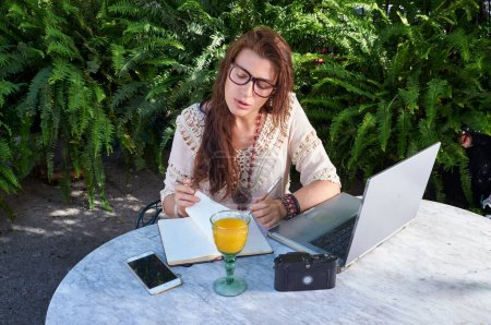 Téléchargez les photos : Une femme en lunettes travaille sur un ordinateur portable dans un parc, profitant de la liberté et de la beauté de la nature. Elle combine parfaitement travail et loisirs. - en image libre de droit