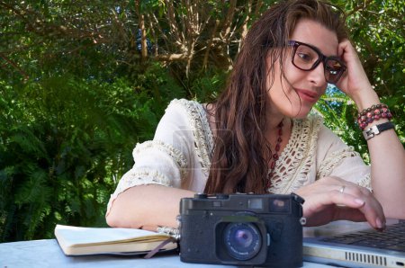 Photo for A woman with eyeglasses and retro hairstyle sits outdoors with a laptop and camera, surrounded by nature. She is studying or working on digital projects, embracing bleisure activities. - Royalty Free Image