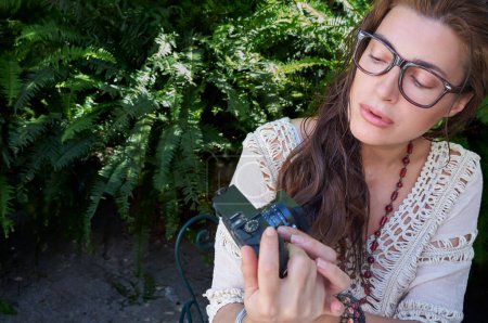 Photo for A woman in casual clothing, with long brown hair and eyeglasses, works on her vintage canera outdoors surrounded by lush greenery, studying and enjoying her job. - Royalty Free Image