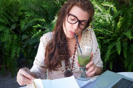 Photo for A young woman in glasses sits outdoors, working on a laptop and drinking a green juice. She studies and communicates digitally, surrounded by nature. A perfect picture of remote work and education. - Royalty Free Image