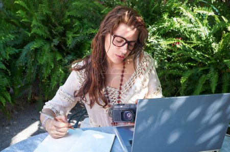 Foto de Una joven con cabello castaño largo y gafas trabaja al aire libre, mezclando tecnología y naturaleza. Estudiar y trabajar, abrazar el estilo de vida digital nómada. - Imagen libre de derechos