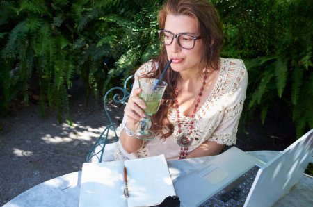 Photo for A happy nomad worker studies outside in spring with a laptop and wears casual clothes and glasses. Technology connects her with work and nature. - Royalty Free Image