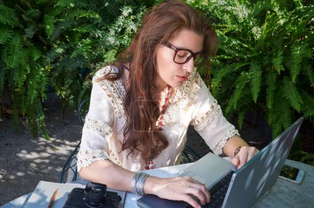 Photo for A woman with brown hair enjoys working outdoors on her laptop, combining work and leisure seamlessly. She wears glasses and casual clothes, embracing the freedom of being a digital nomad. - Royalty Free Image