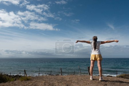 Photo for A confident woman stands on the shore, arms outstretched towards the sky, in a contemplative moment of self-expression and self-care amidst the calming beach and ocean scenery. - Royalty Free Image