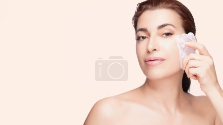 Téléchargez les photos : Une femme élégante utilise un outil gua sha en quartz rose sur sa peau sur un fond rose doux, incarnant la sérénité et l'élégance des soins de la peau. Espace de copie - en image libre de droit