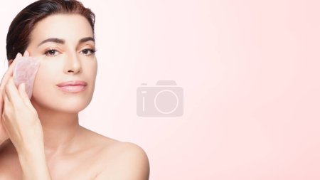 Téléchargez les photos : Une femme gracieuse s'engage dans une routine de soins de la peau en utilisant un gua sha en quartz rose sur un fond rose doux, soulignant la beauté et le bien-être. - en image libre de droit