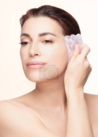 Téléchargez les photos : Une femme sereine applique une pierre gua sha en quartz rose sur sa joue pour des soins de la peau et un soin de beauté relaxant dans un cadre de studio. - en image libre de droit