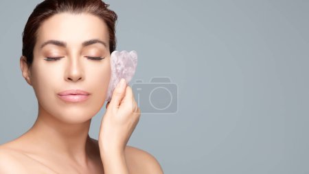 Téléchargez les photos : Une jolie femme brune utilisant une pierre Gua Sha en quartz rose sur son visage pour des soins de la peau et un traitement anti-âge sur un fond bleu doux. - en image libre de droit