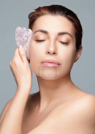 Graceful middle-aged woman performing a gua sha facial massage. This spa ritual focuses on anti-aging and skin rejuvenation. Ideal for beauty tutorials and wellness blogs. Isolated on grey.