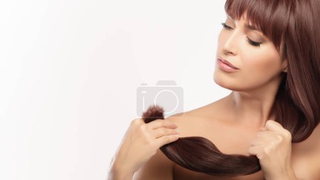 Téléchargez les photos : Portrait d'une jolie femme tenant ses longs cheveux bruns brillants, mettant l'accent sur la beauté et les soins capillaires sur un fond blanc isolé avec espace de copie. - en image libre de droit