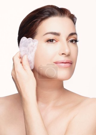 Eine schöne Frau demonstriert die Verwendung eines rosafarbenen Gua-Sha-Steins auf ihrem Gesicht, der eine gesunde Hautpflege fördert und Wohlbefinden ausstrahlt. Vertikales Format