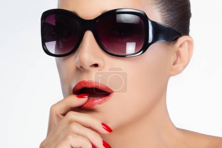 A fashionable and confident woman with large black sunglasses and vibrant red lipstick and nails poses elegantly, isolated on a white background with copy space.