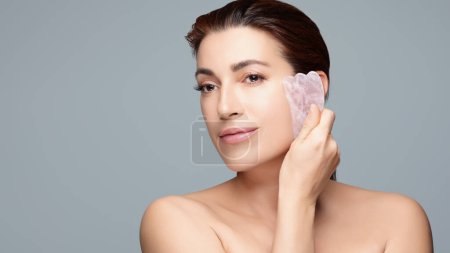 Mature woman performing facial massage with a gua sha stone, isolated on a light grey background, conveying concepts of beauty, skincare, and anti-aging.