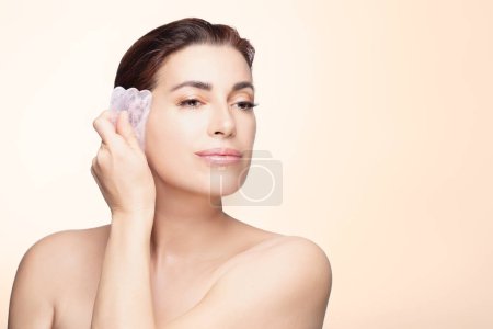 A serene woman gently uses a gua sha stone on her face, promoting skincare and relaxation in a soothing pastel backdrop.