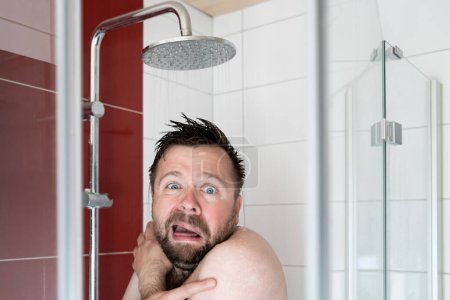 European man takes a shower under cold water, he freezes and looks miserable. Concept of savings and energy crisis. 