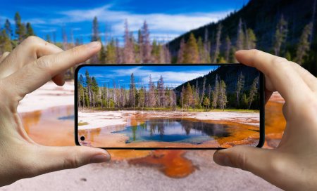 Téléchargez les photos : Touriste prenant une photo avec un téléphone portable d'une source chaude dans le parc national de Yellowstone, États-Unis. - en image libre de droit