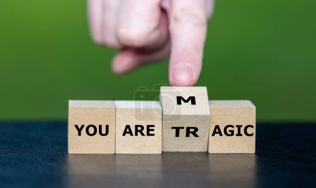 Hand turns wooden cube and changes the expression ' you are tragic' to 'you are magic'.