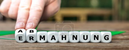 Hand turns dice and changes the German word 'Ermahnung' (admonition) to 'Abmahnung' (dissuasion).