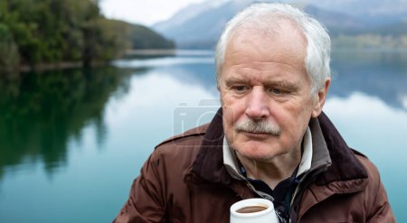 Thoughtful senior man drinking coffee alone at a mountain lake. 
