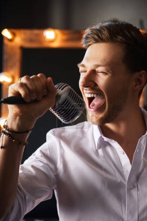 Photo for Young handsome bearded man is singing loudly into a microphone with his eyes closed at home with a makeup mirror behind his back. - Royalty Free Image