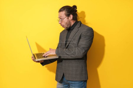 Foto de Sorprendido hombre barbudo con gafas está utilizando ordenador portátil sobre fondo amarillo - Imagen libre de derechos