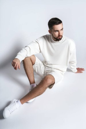 Photo for Handsome man wearing white sweatshirt and shorts with wireless earbuds sitting against gray background - Royalty Free Image
