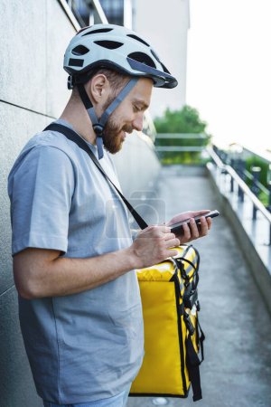 Téléchargez les photos : Jeune messager express livraison de nourriture avec sac isolé sur le côté regarde son téléphone. - en image libre de droit